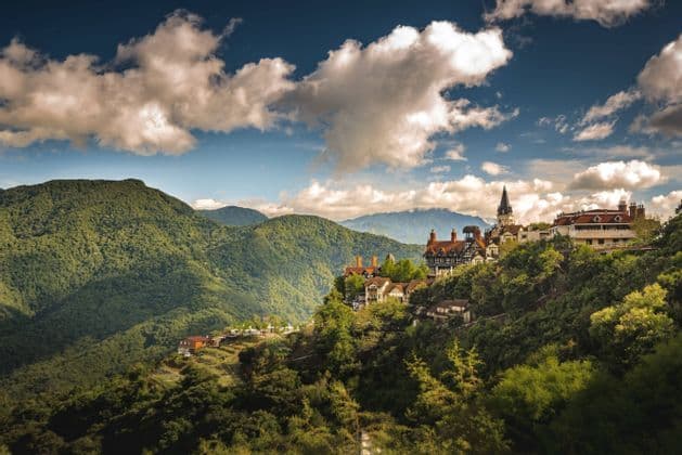 A European-style castle and village nestled in lush green mountains under a partly cloudy sky.