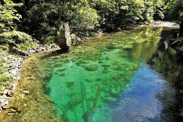 Un fiume limpido e turchese scorre attraverso una fitta foresta, con un'antica colonna di pietra che si erge sulla sponda rocciosa.