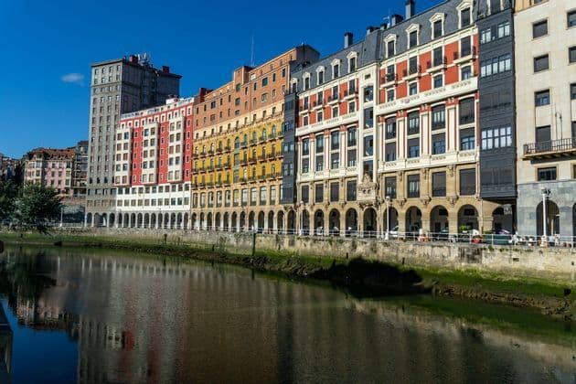 Una fila di edifici colorati a più piani con portici ad arco si erge lungo un fiume, le cui riflessioni si specchiano nell'acqua.