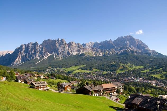 Un village de montagne ensoleillé avec des maisons en bois dispersées dans une vallée verdoyante, dominé en arrière-plan par une chaîne de montagnes déchiquetée et rocheuse.