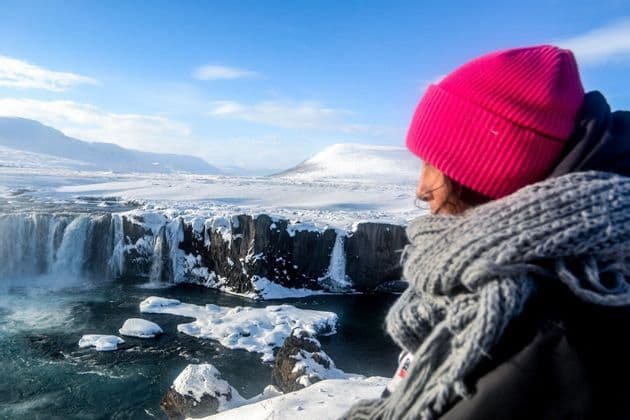 Une personne portant un bonnet rose et une écharpe grise contemple une grande cascade partiellement gelée dans un vaste paysage enneigé sous un ciel bleu clair.