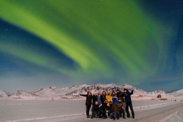Eine WeRoad-Gruppe posiert für ein Foto unter den grünen Nordlichtern in einer verschneiten Berglandschaft.