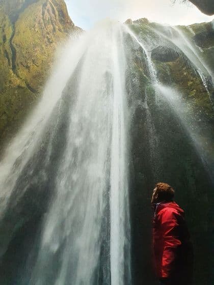 Una persona con una giacca rossa si erge ai piedi di un'imponente cascata, ammirando l'acqua che precipita da una scogliera muschiosa.