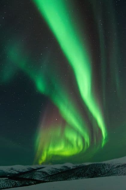 Helle grüne Bänder des Nordlichts leuchten am sternenklaren Nachthimmel über einer verschneiten Bergkette.
