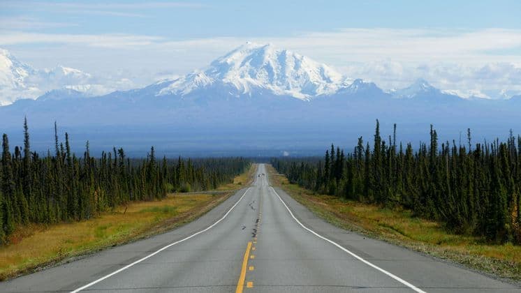 A straight paved road stretches through a forest of evergreens towards a large, snow-capped mountain range in the distance.