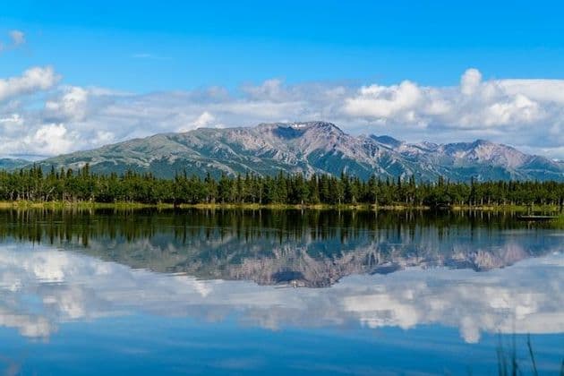 A mountain range and a line of trees reflecting on the calm surface of a lake under a partly cloudy sky.