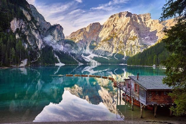 Una casa de botes de madera sobre pilotes y una fila de botes de remo en un lago tranquilo de color turquesa, con grandes montañas y un cielo azul reflejados en el agua.