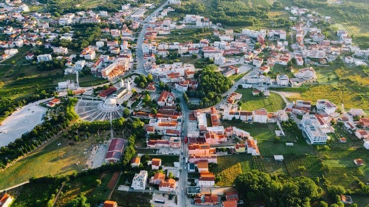 Vista aerea di un vasto paese con edifici dai tetti rossi e una grande piazza circolare, circondato da rigogliosi campi verdi e alberi.
