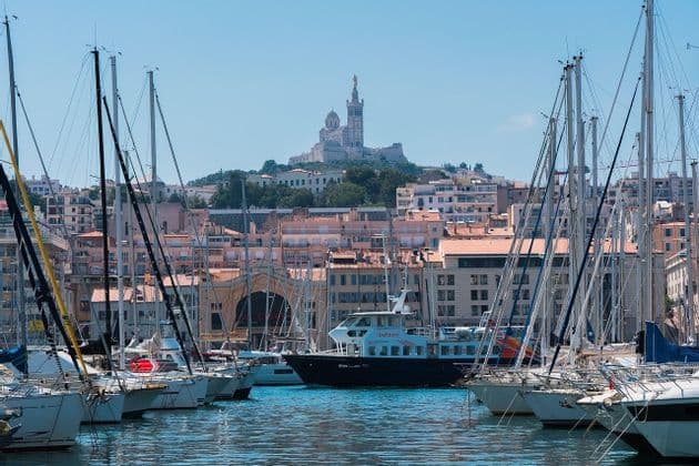 A view of a harbor filled with docked sailboats, with a bustling city and a large basilica on a hill in the background under a clear blue sky.
