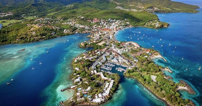 Une vue aérienne d'une ville côtière sur une presqu'île verdoyante, avec un port de plaisance et des voiliers dans la mer d'un bleu et turquoise limpide.