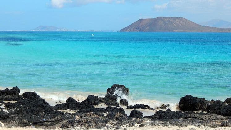 Las olas rompen contra rocas volcánicas negras en una playa de arena, con agua turquesa y una gran isla al fondo.