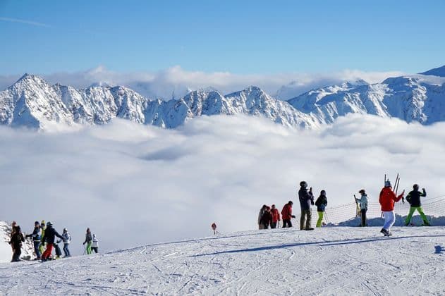 Viaje en grupo WeRoad de esquiadores en la cima de una montaña nevada, observando picos sobre un mar de nubes bajo un cielo azul.