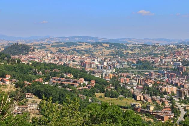 Una veduta dall'alto di una vasta città con tetti rossi, incastonata tra lussureggianti colline verdi sotto un limpido cielo azzurro.