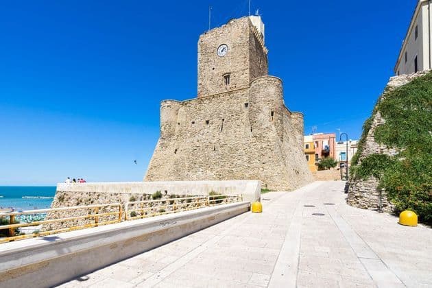Un imponente castello in pietra con torre dell'orologio si erge vicino al mare sotto un cielo azzurro e limpido in una giornata di sole.