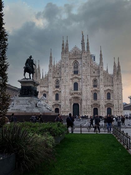 Una ornamentada catedral gótica vista desde una plaza con una estatua ecuestre y gente paseando bajo un cielo nublado.