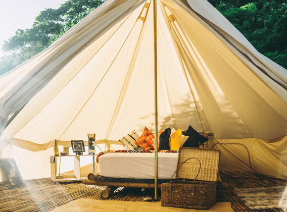 L'interno illuminato dal sole di un'ampia tenda glamping con un letto di bambù, cuscini colorati e un cesto di vimini su un pavimento di legno.
