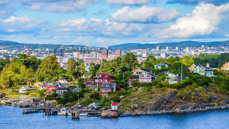 Colorful houses dot a green, rocky shoreline in front of a distant city skyline under a partly cloudy sky.