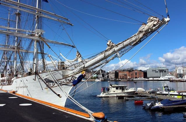 A large, white, multi-masted sailing ship is docked in a city harbor under a clear blue sky.