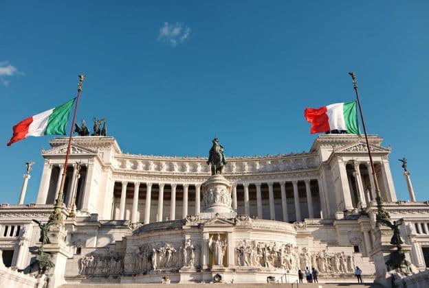 Due bandiere italiane sventolano sopra un grande monumento in marmo bianco con colonne e una statua equestre sotto un cielo azzurro chiaro.