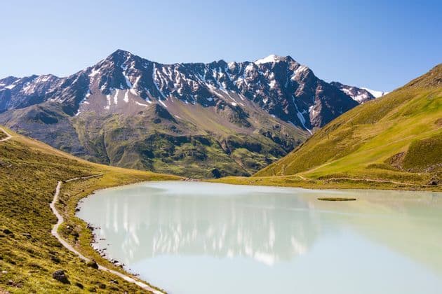 Ein ruhiger, milchiger Alpensee spiegelt an einem sonnigen Tag die schneebedeckten Gipfel einer Bergkette in einem grünen Tal.