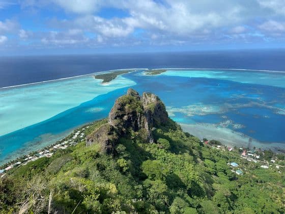 Una vista dall'alto di una lussureggiante cima montuosa verde che si affaccia su un'isola tropicale con una laguna turchese e barriere coralline.