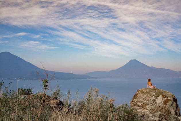 Una persona con un cappello è seduta su una grande roccia, ammirando un vasto lago e montagne lontane sotto un cielo azzurro nuvoloso.