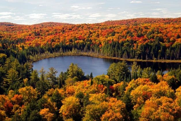 Un lago blu è circondato da una fitta foresta con fogliame autunnale arancione e giallo che copre dolci colline sotto un cielo parzialmente nuvoloso.