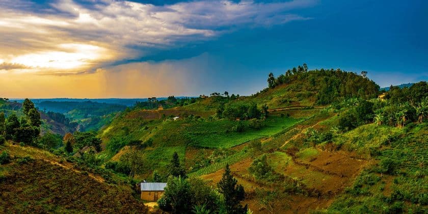 Une vaste étendue de collines verdoyantes et ondulantes, parsemée de terrasses agricoles et de quelques petites maisons, sous un ciel partagé entre soleil et nuages.