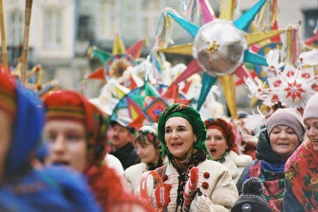 Eine Frau in traditioneller Kleidung mit einem grünen Kopftuch lächelt und hält ein Tamburin während einer festlichen Straßenparade mit bunten Sternen.