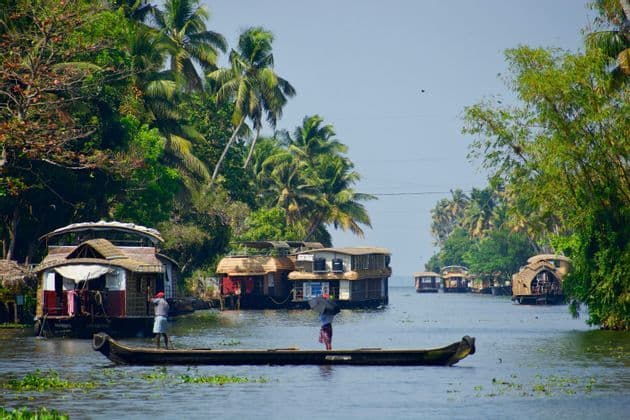 Due persone in una piccola canoa remano su un fiume tranquillo, superando diverse case galleggianti tradizionali ormeggiate lungo una riva costeggiata da palme rigogliose.
