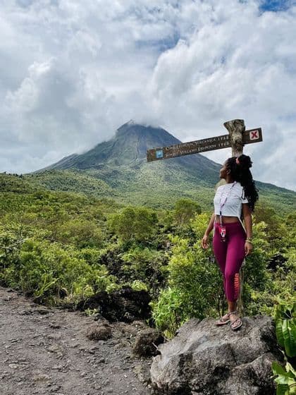 Una mujer en una ruta de senderismo se para en una roca, mirando hacia un gran volcán parcialmente cubierto por nubes.