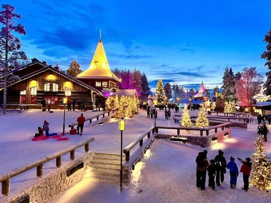 Un pueblo nevado al anochecer con gente caminando entre cabañas de madera iluminadas y árboles de Navidad decorados.