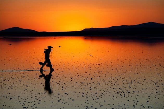 Una sagoma con cappello conico e bilanciere in spalla cammina in acque basse e riflettenti al tramonto.