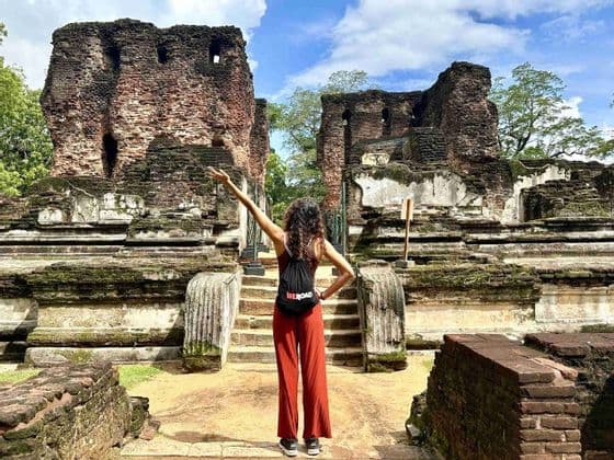 Une femme vue de dos, portant un sac à dos WeRoad, se tient devant d'anciennes ruines de briques couvertes de mousse, un bras levé.