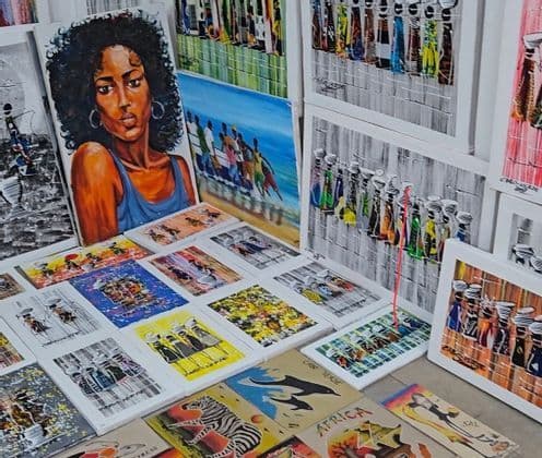 Eine Sammlung farbenfroher Leinwandbilder, darunter ein Frauenporträt, die afrikanische Motive zeigen und an einem Marktstand präsentiert werden.