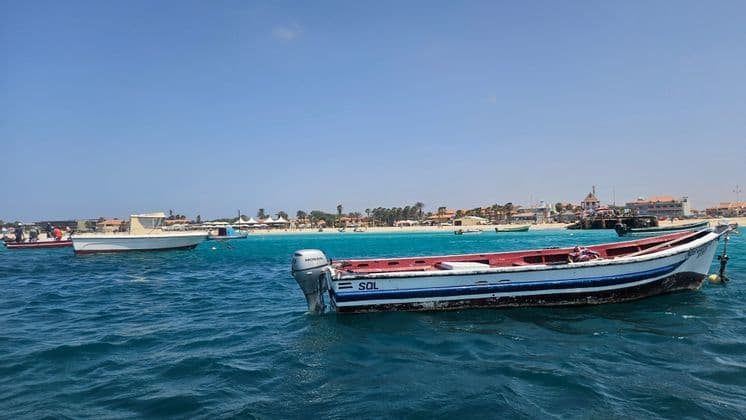 Ein rot-weißes Motorboot schwimmt auf blauem Wasser, mit einem Sandstrand und einer Küstenstadt im Hintergrund.