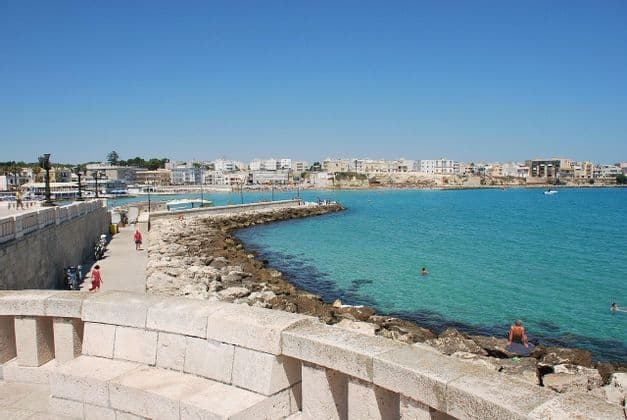 Vista soleggiata di una località costiera con un lungomare in pietra, un frangiflutti roccioso e bagnanti nel mare turchese.