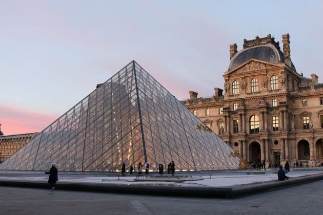 Una gran pirámide de cristal en un patio de piedra con un edificio histórico ornamentado al fondo al anochecer.