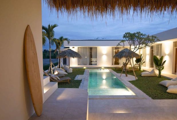 Un moderno cortile di villa al tramonto, con piscina illuminata, ombrelloni in paglia e una tavola da surf appoggiata al muro.