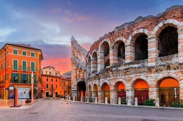 Un anfiteatro in pietra si trova accanto a un edificio arancione, su una strada di ciottoli, sotto un cielo colorato al tramonto.