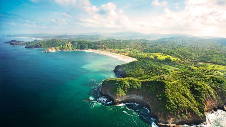Una veduta aerea di una costa dove rigogliose colline verdi incontrano un oceano turchese, con una spiaggia di sabbia che si estende lungo una baia lontana.