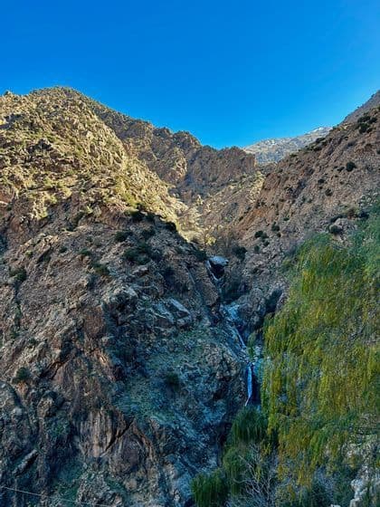 Una cascada fluye por un desfiladero rocoso entre laderas de montañas soleadas bajo un cielo azul claro.