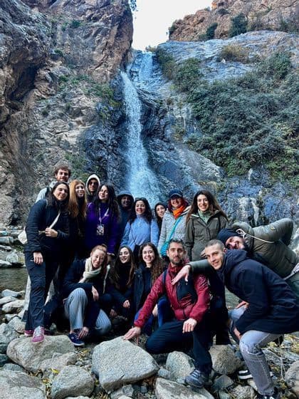 Un viaje en grupo de WeRoad posando para una foto en rocas en la base de una cascada que cae por un acantilado empinado.