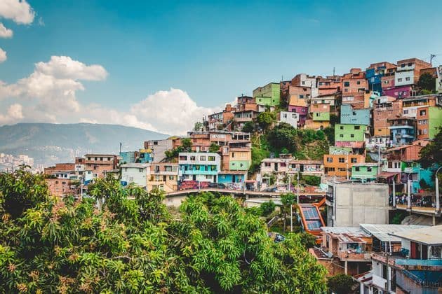 A dense neighborhood of colorful houses covers a green hillside, with an outdoor escalator and distant mountains under a blue sky.