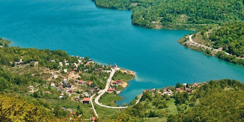 Eine Luftaufnahme eines kleinen Dorfes am Ufer eines hellblauen Sees, eingebettet in dichte, grüne, bewaldete Hügel.