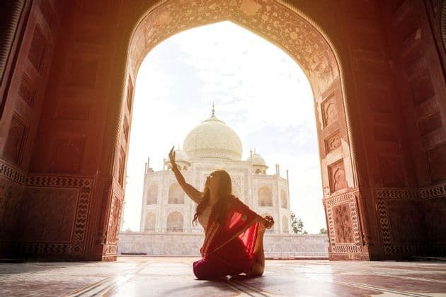 Una donna in un sari rosso esegue una posa yoga sul pavimento all'interno di un grande arco, che incornicia la vista di un mausoleo di marmo bianco.