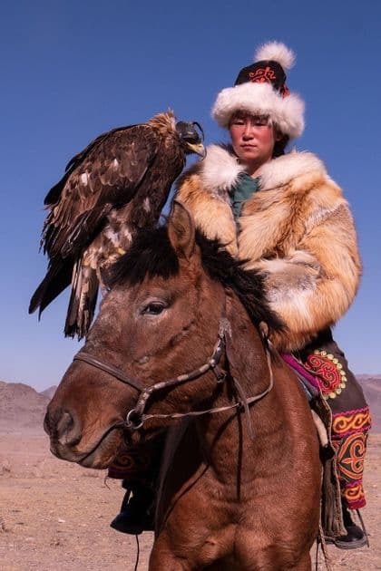 Una persona in tradizionale cappotto e cappello di pelliccia siede su un cavallo marrone, con una grande aquila incappucciata appollaiata sul braccio.