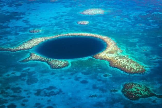 An aerial view of a large, deep blue marine sinkhole encircled by a coral atoll in shallow turquoise ocean water.