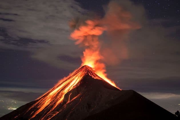A volcano erupts at night, with glowing lava flowing down its dark slopes under a starry sky.