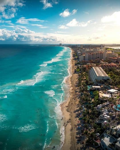 An aerial view of a crowded sandy beach with turquoise water, lined with hotels and palm trees under a blue sky with white clouds.